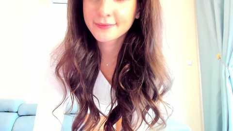 Media: Video of a smiling young woman with long, wavy brown hair, wearing a white top, standing indoors near a blue couch and light-colored walls.