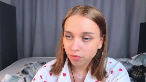 Media: Video of a young, fair-skinned woman with straight, shoulder-length blonde hair, wearing a white shirt with red heart patterns and large hoop earrings. She has a neutral expression, sitting in a bedroom with grey curtains and a bed with patterned sheets.