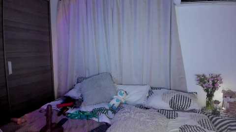 Media: Video of a cluttered bedroom with white bedspread, pillows, and a stuffed animal, surrounded by a dark wooden wardrobe, white curtains, and a vase of flowers on a nightstand.