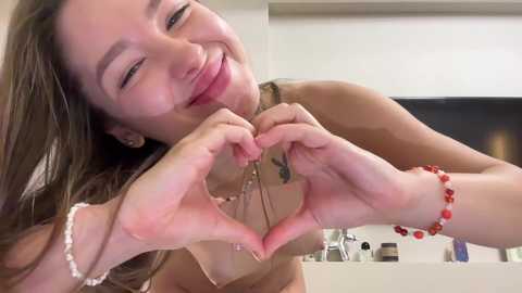 Media: Video of a smiling, light-skinned woman with long brown hair, wearing a red beaded bracelet, forming a heart shape with her hands in front of a blurred background.