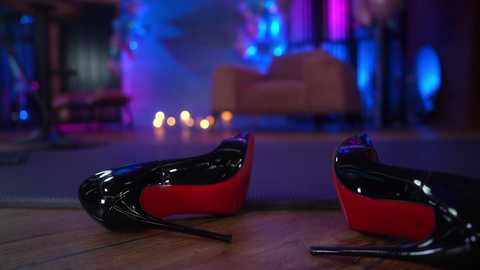 Media: Video of black patent leather high heels with red soles lying on a wooden floor, blurred background featuring a plush armchair, dim lighting, and soft blue and purple ambient lighting.