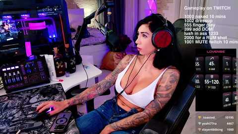 Media: A tattooed woman in a white bra and jeans sits in a gaming chair, wearing headphones, using a gaming mouse on a desk with monitors and equipment.