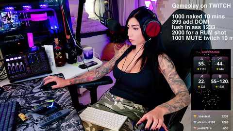 Media: Video of a tattooed, pale-skinned woman with long black hair, wearing a tight black tank top and camouflage pants, gaming in a dimly lit room with colorful lights and gaming gear.