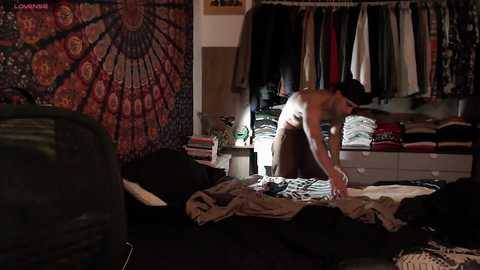 Media: Video of a messy, dimly lit bedroom with a bare-chested man folding clothes on a bed, surrounded by scattered clothes and a tapestry on the wall.