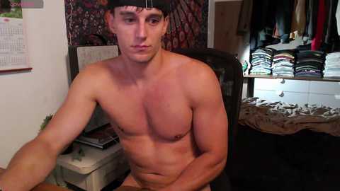 Media: Video of a shirtless, muscular man with a black cap, sitting at a desk in a cluttered room. Background shows a calendar, clothes, and a messy bed.