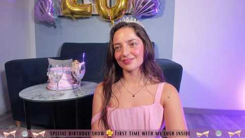 Media: Video of a smiling young woman with long dark hair, wearing a pink dress and silver tiara, sitting in a room with a dark couch, gold and silver balloons, and a birthday cake on a round table.