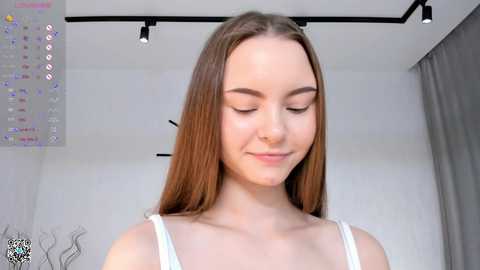 Media: Video of a fair-skinned, young woman with long, straight brown hair, wearing a white spaghetti-strap top, smiling softly. Background features a white wall with a colorful, digital art piece and modern, black track lighting.