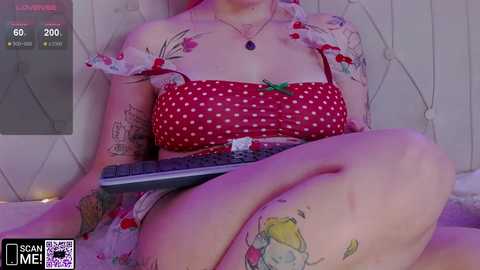 Media: Video of a fair-skinned woman with tattoos, wearing a red polka-dot dress, sitting on a bed, holding a remote control.