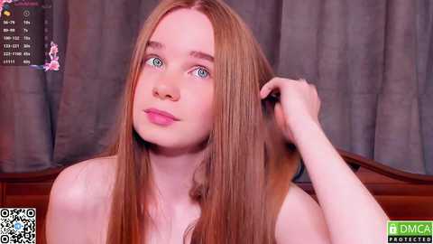 Media: Video of a fair-skinned, young woman with long, straight red hair and blue eyes, touching her hair. She is topless, with a soft smile. Background features grey curtains and a wooden headboard.