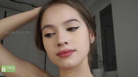 Media: Video of a young woman with fair skin, brown eyes, and straight brown hair. She has a slim physique and wears a subtle makeup look with winged eyeliner and light pink lipstick. She is indoors, likely in a bathroom, with a white sink and mirror in the background.