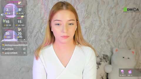 Media: Video of a young, fair-skinned, blonde woman with straight hair wearing a white V-neck top, sitting in a cozy room with a textured wall and a plush bear.