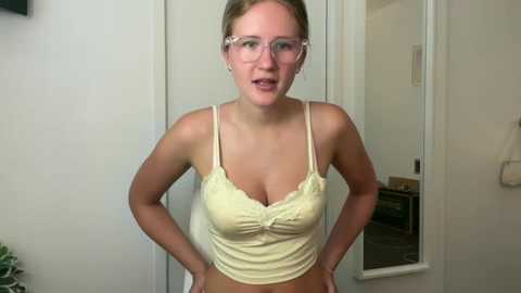 Media: Video of a light-skinned, blonde woman with glasses wearing a yellow lace-trimmed camisole, hands on hips, in a minimalistic, white-walled room.