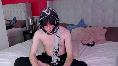 Media: A video of a shirtless young man with pale skin, black hair, and a black-and-white scarf, sitting on a bed with white sheets, pink pillows, and a tufted headboard.