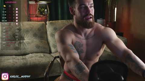 Media: Video of a shirtless, muscular man with tattoos, a beard, and a serious expression, sitting on a beige couch in a dimly lit room, holding a black garment.