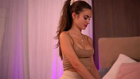 Video of a young, slender, fair-skinned woman with long brown hair in a ponytail, wearing a beige tank top and shorts, standing indoors next to a beige bed, against a soft purple and white background.