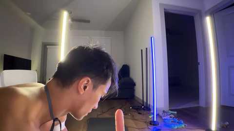 Media: A video of a man with short black hair, shirtless, standing in a dimly lit room with white walls, holding a large pink dildo, surrounded by blue light sticks and cleaning supplies.