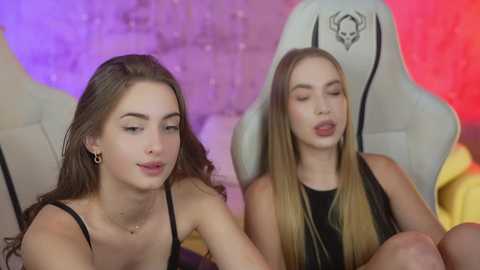 Media: Video of two young women with fair skin, long hair, wearing black tops, seated on gaming chairs. Background features a purple and red gradient wall.