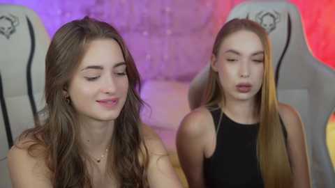 Media: Video of two young women with long hair in a dimly lit, modern indoor setting. One woman with wavy brown hair, wearing a black top, smiles softly. The other, with straight blonde hair, wears a black sleeveless top, looking serene.