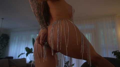 Media: Video of a topless woman with a tattooed arm, wearing a silver, tassel-covered skirt, posing provocatively in a dimly lit room with sheer curtains and potted plants.