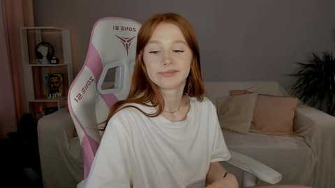 Media: Video of a young, fair-skinned redhead with long hair in a white T-shirt, sitting in a pink gaming chair in a cozy, modern living room.