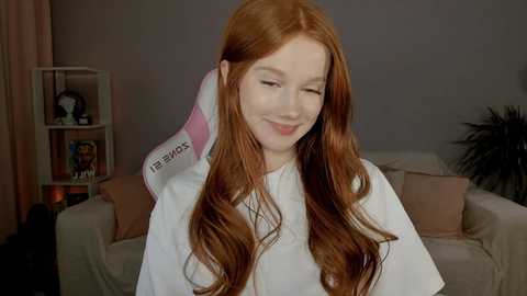 Media: Video of a smiling young woman with long red hair, wearing a white beauty cape, in a cozy living room with soft lighting, a beige sofa, and a bookshelf.