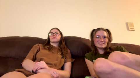 Media: A video of two young women with glasses, sitting side-by-side on a brown couch, wearing casual clothing, in a simple indoor setting.