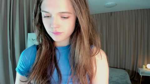 Media: A video of a young woman with long, straight brown hair, wearing a blue top, standing indoors. She has a contemplative expression. The background features beige curtains and a partially visible bed.