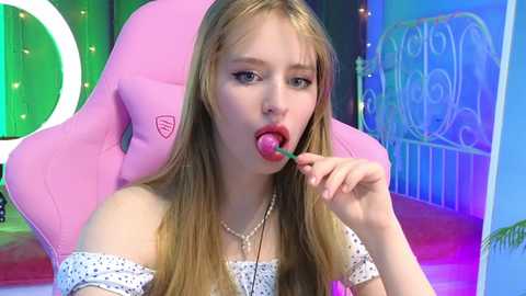 Media: Video of a young, light-skinned woman with long blonde hair licking a strawberry, sitting in a pink gaming chair. She wears a white off-shoulder top with polka dots. Background features a colorful, cartoonish room with a metal bed frame.