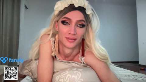 Media: Video of a fair-skinned, blonde woman in a maid outfit with long hair, large earrings, and dramatic makeup, lying on a bed with a beige floral sheet in a simple room.