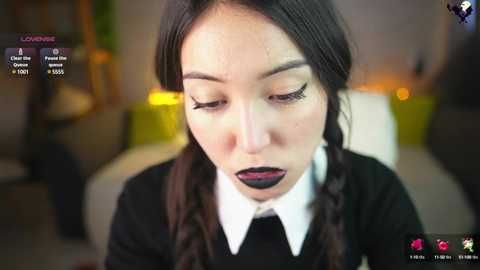 Media: Video of an Asian woman with dark hair in braids, wearing a black sweater over a white collared shirt, makeup with dark lipstick, and a green background.