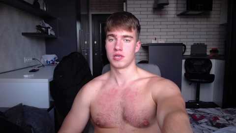 Media: Video of a shirtless young white man with short brown hair, sitting in a modern, dimly-lit apartment with a white brick wall, kitchen, and messy living area.