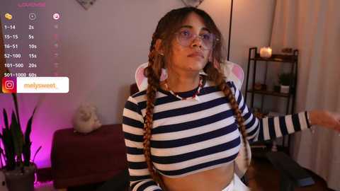 Media: A video of a young woman with light brown skin, wearing glasses and a striped crop top, sitting in a dimly lit room.