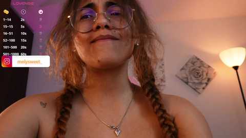 Media: Video of a smiling young woman with glasses, wavy brown hair, and a tattoo, wearing a heart necklace, in a softly lit room with a white wall and a rose print.