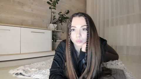 Media: Video of a young Caucasian woman with long, straight, dark brown hair, wearing a black jacket, lying on a white shag rug in a modern, minimalist room with light wood walls, white furniture, and a potted plant.