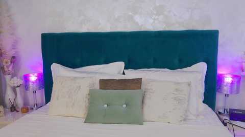 Media: Video of a modern bedroom with a teal tufted headboard, white pillows, a green cushion, purple bedside lamps, and a textured white wall.