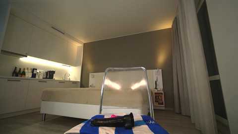 Media: Video of a modern, minimalist bedroom with a beige bed, white cabinets, and a small kitchenette. A striped beach towel and a black shoe are prominently displayed on the floor.