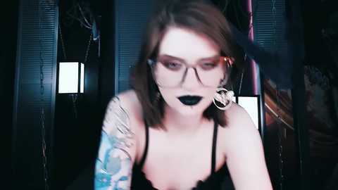 Media: Video of a pale-skinned woman with shoulder-length brown hair, wearing black glasses, black lipstick, and a black lace top, sporting tattoos on her arm. She is in a dimly lit room with dark walls and chain curtains.