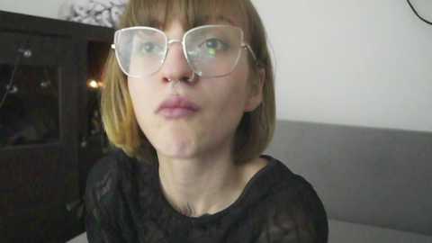 Media: Video of a fair-skinned woman with shoulder-length blonde hair, wearing large, round glasses, a black sheer top, and a visible nose ring. She puckers her lips. Background shows a dark wooden cabinet with glass doors.