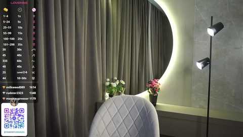 Media: Video of a modern, dimly lit bedroom featuring a round mirror with a bright white light, gray quilted chair, and gray curtains.