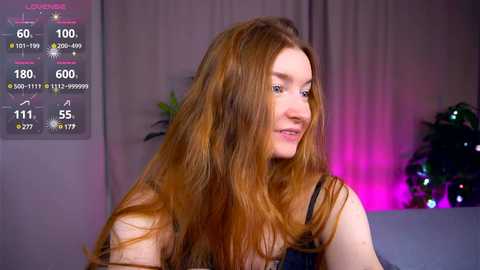 Media: Video of a young Caucasian woman with long, straight red hair, wearing a sleeveless black top. Background includes a digital screen displaying health statistics and a potted plant.