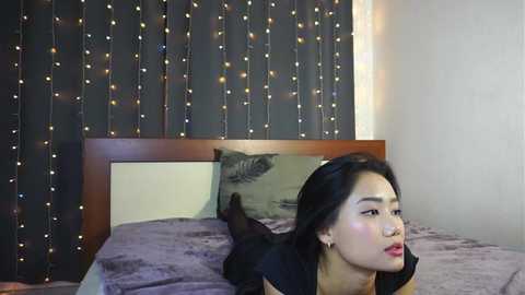 Media: Video of an East Asian woman with long black hair, lying on a bed with a dark purple blanket, wearing black clothing. Background features a wooden headboard and string lights.