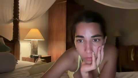 Media: A video of a woman with dark hair and olive skin, lying on a bed, wearing a yellow top, looking tired, in a dimly lit bedroom with wooden furniture.