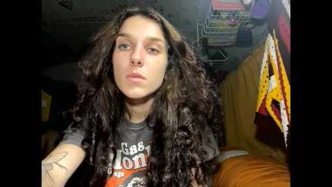 Media: A video of a young person with long, curly dark hair, wearing a gray T-shirt with a graphic design. The background shows a cluttered room with a bed, storage shelves, and yellow and orange clothing items.