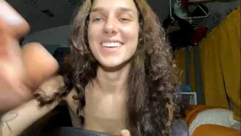 Media: Video of a young woman with long, curly brown hair, light skin, and a nose ring, smiling. Background shows a cluttered room with yellow curtains, a ceiling fan, and a messy bed.