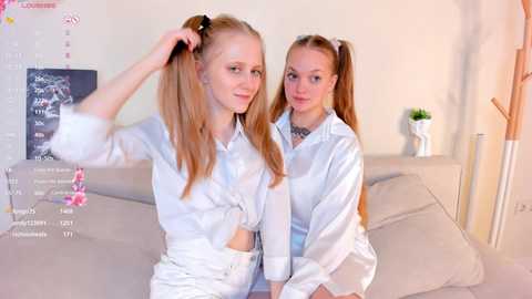 Media: Video of two young, slim Caucasian women with long, light brown hair tied in pigtails, wearing white silk shirts and high-waisted pants, sitting on a beige couch in a minimalist room.