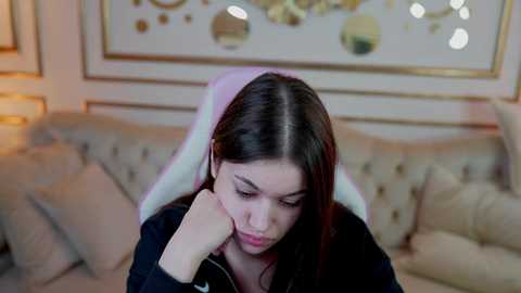 Media: Video of a young woman with long dark hair, wearing a black hoodie, sitting on a white gaming chair in a cozy, beige-lit living room with decorative framed art and soft cushions in the background.