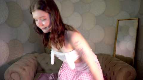 Media: A video of a young woman with long, wavy brown hair, wearing a white strapless top and pink plaid skirt, bending forward in a dimly lit, beige-tufted sofa room with circular wallpaper.