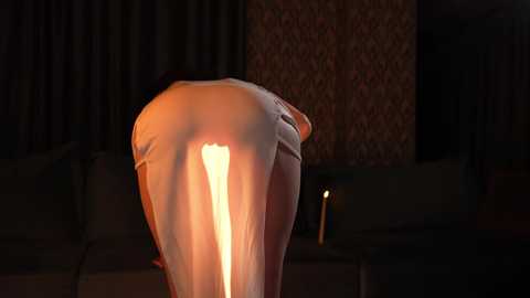 Media: A video captures a close-up of a person bending over, illuminated by a candle, revealing their buttocks and a partially visible, lit candle on a table in a dimly lit room with patterned wallpaper and dark curtains.