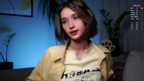 Media: Video of a young Caucasian girl with shoulder-length brown hair, wearing a yellow graphic T-shirt and open shirt, smiling in a dimly-lit living room with green plants.