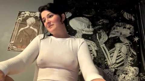 Media: Video of a young woman with pale skin, dark hair, and a white ribbed top, standing in front of a black tapestry with a detailed skeleton design.